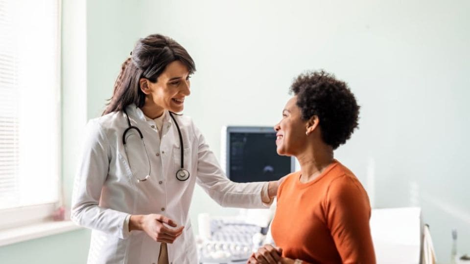 A doctor with a stethoscope around her neck smiles and talks with a patient in an examination room, seamlessly accessing the hospital management system to ensure efficient and accurate care.