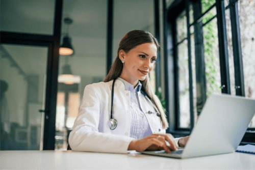 A doctor in a white coat is consulting with a patient via laptop at a desk in a well-lit office with large windows. A doctor in a white coat is consulting with a patient via laptop at a desk in a well-lit office with large windows.